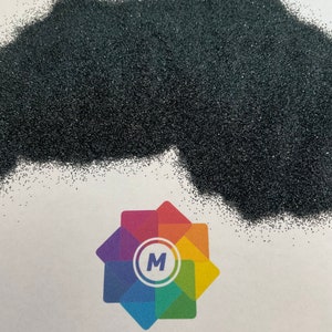 Op de afbeelding: Een close-up van een hoop zwarte mica glitter. De glitter is erg fijn en heeft een lichte glans. De glitter ligt op een wit oppervlak met een kleurrijk logo dat "Mica Man" zegt.