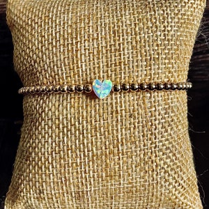 May include: A gold beaded bracelet with a heart-shaped opal charm. The bracelet is on a brown burlap background.