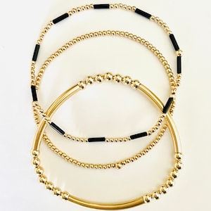 May include: Three gold-coloured bracelets. One bracelet features small gold beads and black cylindrical beads. Another bracelet has small gold beads. The third bracelet has a curved gold tube design with gold beads.