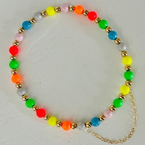 Może przedstawiać: Kolorowy naszyjnik z koralików z naprzemiennymi koralikami neonowymi i szarymi, połączonymi złotymi przekładkami. Naszyjnik ma złoty łańcuszek przymocowany do jednego końca.