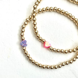 May include: Two gold beaded bracelets with heart-shaped opal charms. One charm is pink and the other is purple.