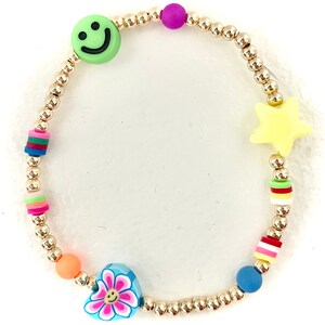 May include: A gold beaded bracelet with colorful charms, including a smiley face, a star, a heart with a flower, and two striped beads.