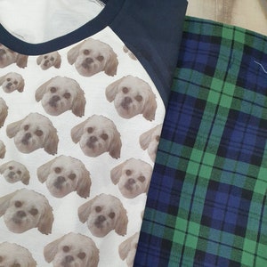 Puede incluir: Una camiseta blanca con una manga raglan azul marino y un patrón de caras de perro repetidas. La camiseta está sobre una tela de cuadros azules y verdes.