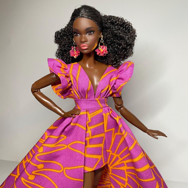May include: A fashion doll with dark brown skin wearing a pink and orange dress with a high low hemline. The doll has curly black hair and is wearing pink flower earrings.