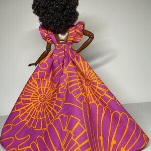 May include: A doll wearing a purple and orange dress with a geometric pattern. The dress has a ruffled neckline and a full skirt.