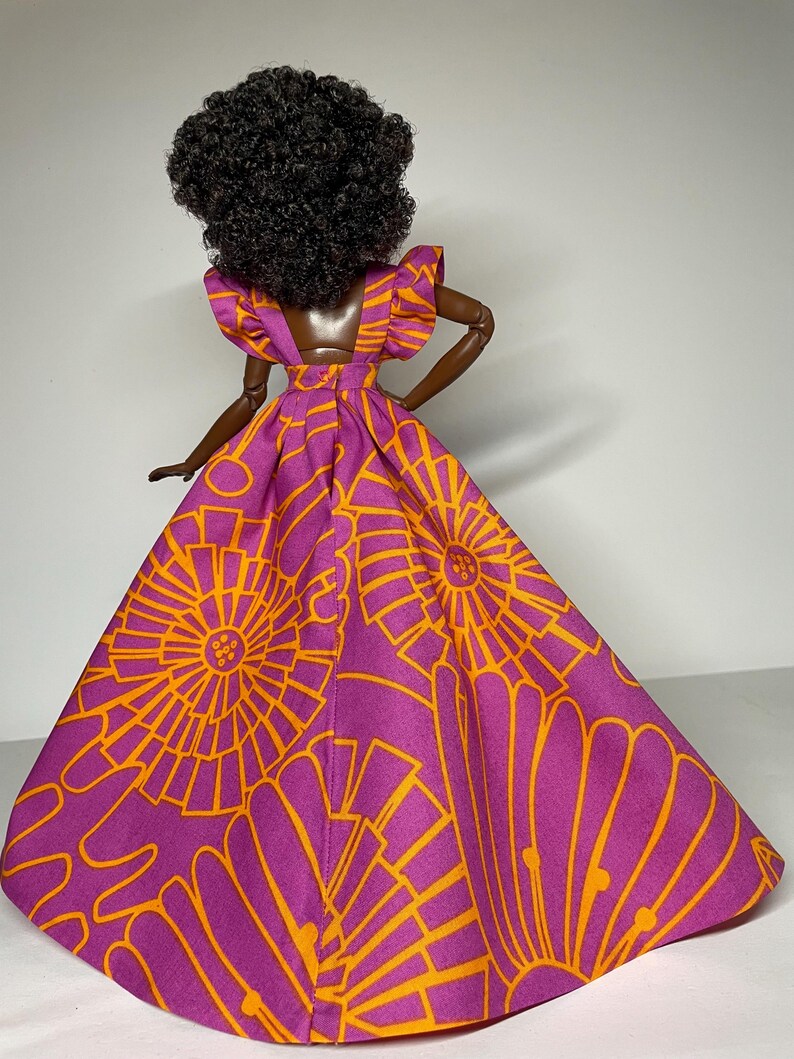 May include: A doll wearing a purple and orange dress with a geometric pattern. The dress has a ruffled neckline and a full skirt.
