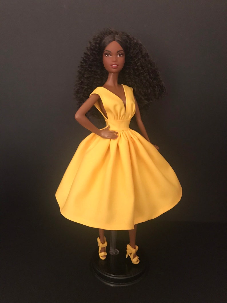 May include: A black doll wearing a yellow sleeveless dress with a V-neckline. The doll has curly black hair and is wearing yellow heels.