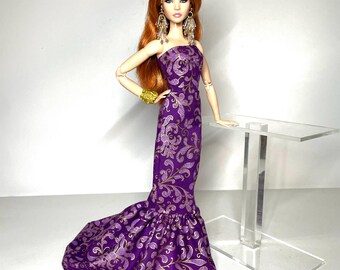 May include: A fashion doll in a purple strapless gown with a gold floral pattern. The doll has long red hair, dangling earrings, and a gold bracelet. The gown has a mermaid-style hemline. The doll is posed next to a clear acrylic table.