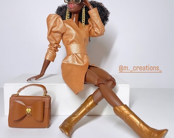 May include: A fashion doll wearing a gold metallic dress with a wrap skirt, gold boots, and gold sunglasses. The doll is sitting on a white surface with a brown leather handbag with a gold bow.