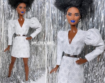 May include: A doll wearing a white dress with a silver belt and a silver background. The doll has blue lipstick and large hoop earrings. The doll's hair is styled in an afro.