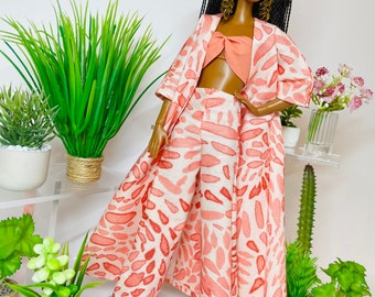 May include: A fashion doll wearing a pink and white patterned pantsuit with a matching kimono-style jacket. The doll has long black braids and is wearing gold earrings.