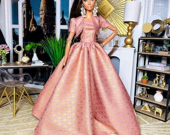 May include: A pink and gold ball gown with a matching cropped jacket. The doll is wearing pearl earrings and has dark brown hair.