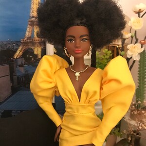 May include: A Black doll with a large afro hairstyle wearing a yellow dress with puffed sleeves and a plunging neckline. The doll is wearing gold earrings, a gold necklace with a key pendant, and gold bracelets. The doll is standing in front of a backdrop of the Eiffel Tower.