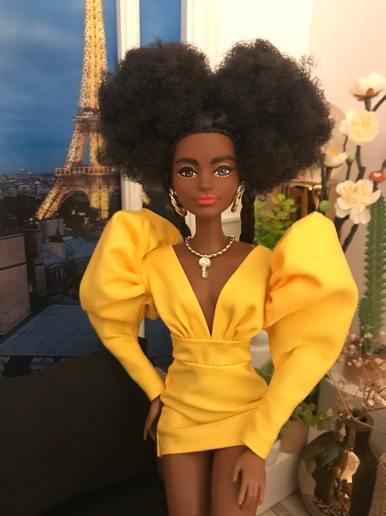 May include: A Black doll with a large afro hairstyle wearing a yellow dress with puffed sleeves and a plunging neckline. The doll is wearing gold earrings, a gold necklace with a key pendant, and gold bracelets. The doll is standing in front of a backdrop of the Eiffel Tower.