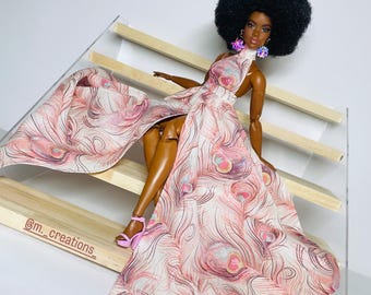 May include: A black doll with an afro hairstyle wearing a pink and white dress with a peacock feather pattern. The doll is sitting on a wooden staircase and wearing pink high heels.