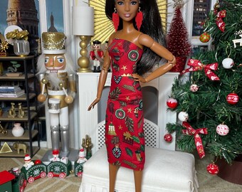 May include: A doll wearing a red, strapless dress with a festive pattern of green leaves, gold stars, and ornaments. The doll has long black hair and red earrings. She is standing in front of a Christmas tree decorated with red bows and ornaments.