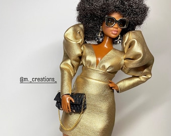 May include: A doll wearing a gold dress with large puffed sleeves and a black purse. The doll has a large afro hairstyle and is wearing sunglasses.