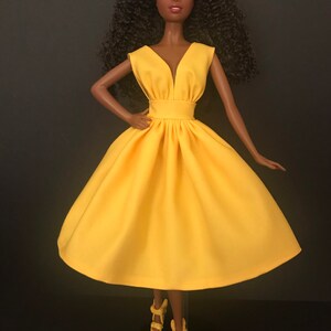 May include: A black doll wearing a bright yellow sleeveless dress with a V-neckline. The doll has curly black hair and is wearing yellow heels.