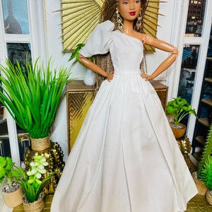 May include: A white one-shoulder gown with a puffed sleeve and a full skirt. The doll is wearing gold earrings and has brown hair.