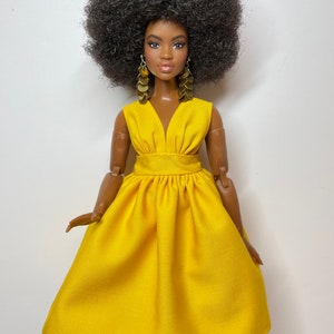 May include: A doll with dark brown skin and a large afro hairstyle is wearing a bright yellow dress with a V-neckline and gold earrings.