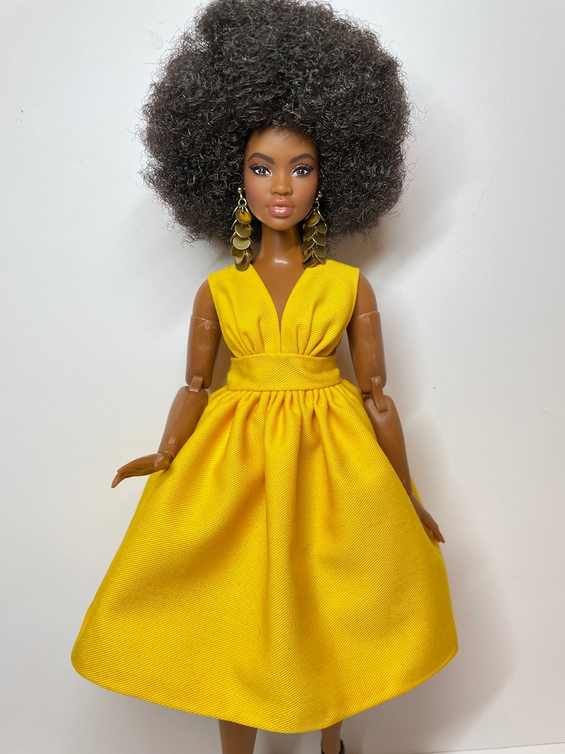 May include: A doll with dark brown skin and a large afro hairstyle is wearing a bright yellow dress with a V-neckline and gold earrings.