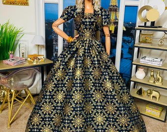May include: A doll wearing a black and gold patterned gown and matching bolero jacket. The dress features a full skirt and a repeating starburst design. The doll has blonde hair and is wearing gold earrings and a bracelet. The background includes a window with a view of the Eiffel Tower.