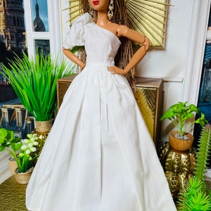 May include: A white one-shoulder dress with a puffed sleeve and a full skirt. The dress is made of a soft, flowing fabric and is perfect for a special occasion. The doll is wearing the dress and has long brown braids.