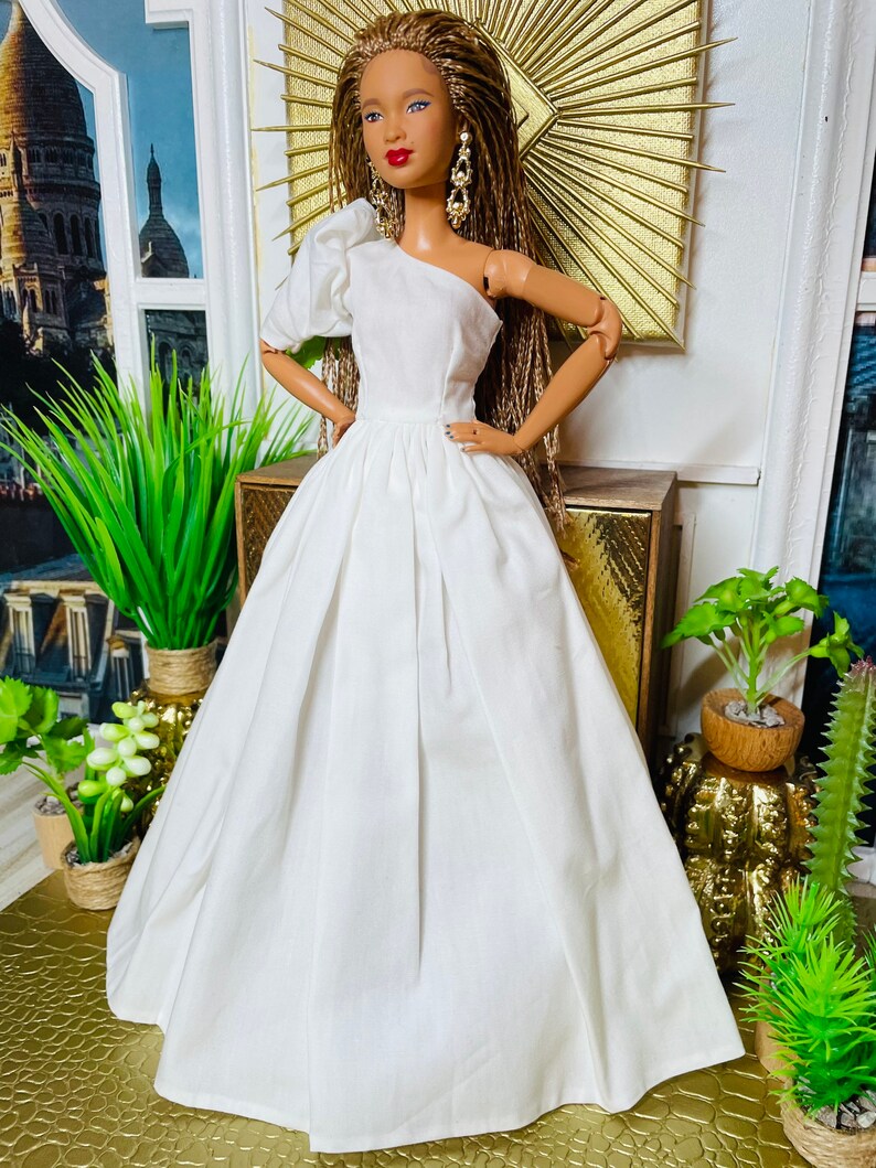 May include: A white one-shoulder dress with a puffed sleeve and a full skirt. The dress is made of a soft, flowing fabric and is perfect for a special occasion. The doll is wearing the dress and has long brown braids.