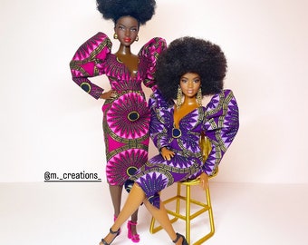 May include: Two Black dolls wearing colorful African print dresses. One doll is wearing a pink dress with gold accents and the other doll is wearing a purple dress with gold accents. Both dolls have black afro wigs and are wearing purple shoes.