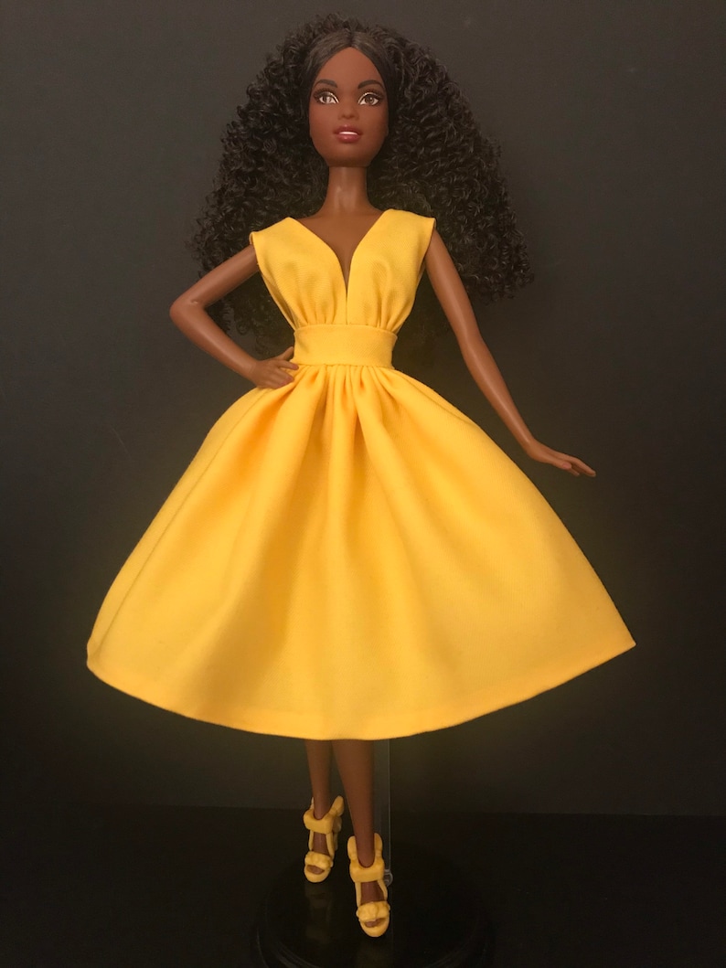 May include: A black doll wearing a bright yellow sleeveless dress with a V-neckline. The dress has a fitted bodice and a full skirt.