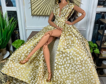 May include: A fashion doll wearing a gold and white patterned gown with a high slit. The doll has long brown hair, brown eyes, and is wearing gold earrings.