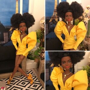 May include: A Black doll with long black braids wearing a yellow dress with large puffed sleeves and a plunging neckline. The doll is wearing gold earrings and a gold necklace with a key pendant. The doll is sitting on a chair with a black cushion. The background is a blurred image of a city skyline.
