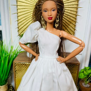 May include: A doll wearing a white one-shoulder dress with a full skirt. The doll has brown braided hair, brown eyes, and red lipstick. She is wearing gold earrings.