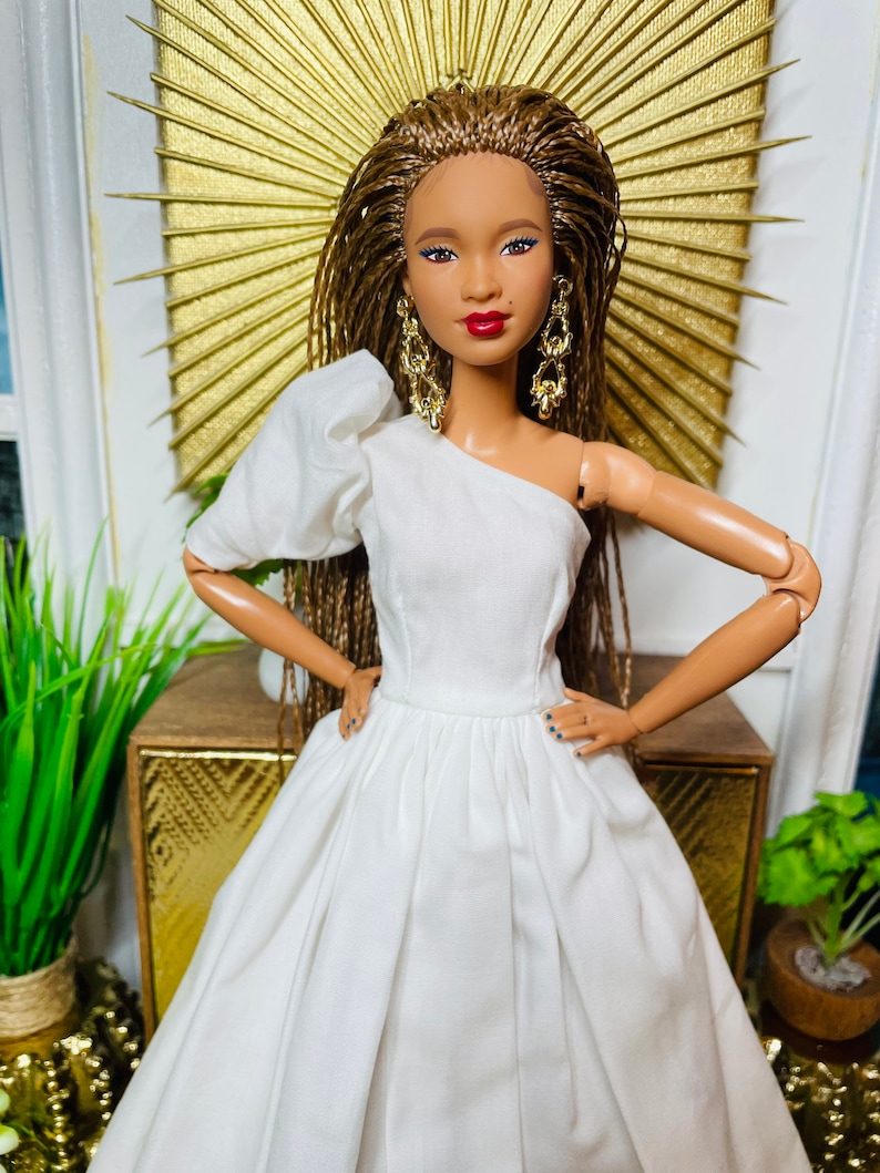 May include: A doll wearing a white one-shoulder dress with a full skirt. The doll has brown braided hair, brown eyes, and red lipstick. She is wearing gold earrings.