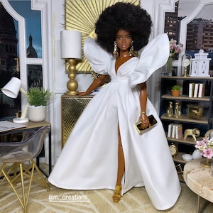 May include: A black doll with an afro hairstyle wearing a white gown with large puffed sleeves and a thigh-high slit. The doll is holding a gold clutch and wearing gold jewelry. The doll is standing in a room with gold accents and a bookshelf.