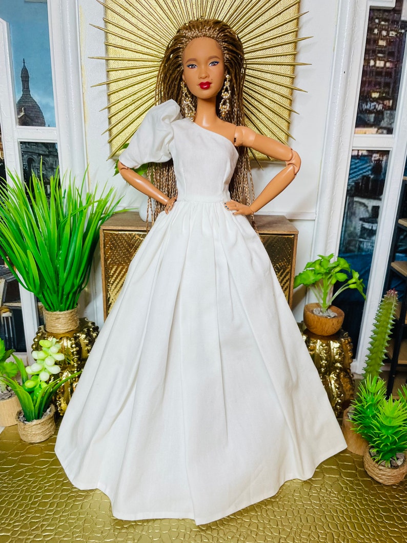 May include: A white one-shoulder gown with a puffed sleeve and a flowing skirt. The doll is wearing the gown and has long brown braids.