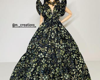 May include: A doll wearing a black gown with a floral pattern in shades of green and gold. The dress has a deep V-neck and ruffled sleeves. The doll has red hair and is wearing gold-toned jewelry.