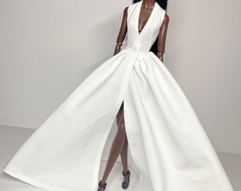 May include: A fashion doll wearing a white sleeveless gown with a plunging neckline and a high slit. The gown has a flowing skirt and is cinched at the waist. The doll has long black hair and dark brown skin.