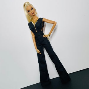 Può includere: Una bambola di moda che indossa una tuta nera con scollatura profonda e una collana d'oro. La bambola ha i capelli biondi raccolti in un'alta coda di cavallo e indossa orecchini d'oro.