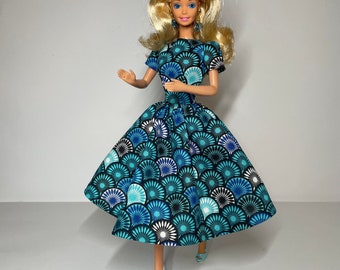 May include: A Barbie doll wearing a blue and white dress with a fan-shaped pattern. The dress has a full skirt and short sleeves. The doll has blonde hair and blue eyes.