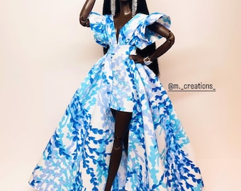 May include: A fashion doll with dark skin wearing a blue and white patterned dress with a high low hemline. The doll has long black hair and is wearing silver earrings. The dress has a plunging neckline and ruffled sleeves. The doll is standing on a white background. @m_creations_