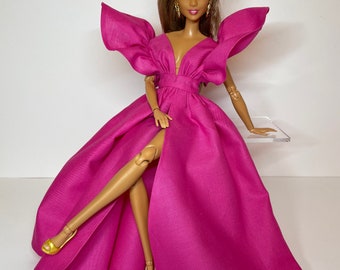 May include: A pink gown with a plunging neckline and large, ruffled sleeves. The gown has a thigh-high slit and is worn by a doll with brown hair and gold shoes.