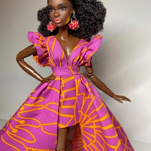 May include: A fashion doll with dark brown skin, wearing a pink and orange dress with a high-low hemline. The doll has curly black hair and is wearing pink flower-shaped earrings.