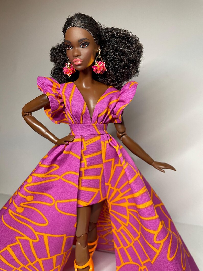 May include: A fashion doll with dark brown skin, wearing a pink and orange dress with a high-low hemline. The doll has curly black hair and is wearing pink flower-shaped earrings.