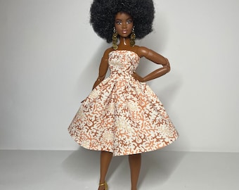 May include: A doll with dark brown skin and an afro hairstyle is wearing a white dress with a coral and brown coral reef pattern. The doll is wearing gold earrings and gold shoes.