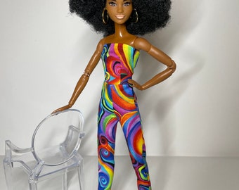 May include: A doll with a large afro hairstyle wearing a colorful jumpsuit and pink shoes. The doll is standing next to a clear plastic chair.