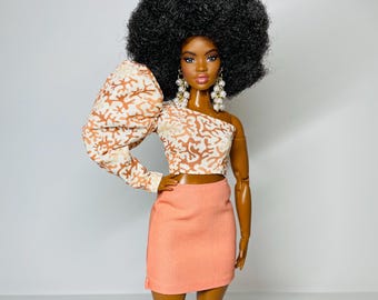 May include: A doll with a large afro hairstyle wearing a peach-colored skirt and a white and brown patterned top with a large puff sleeve.