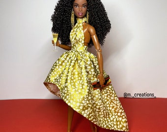 May include: A black doll with curly hair wearing a gold and white patterned dress and gold earrings. The doll is holding a glass of champagne and a small clutch purse.