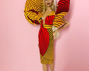 May include: A fashion doll wearing a yellow, red, and black striped dress with a large, ruffled collar. The doll has blonde hair and is wearing gold earrings.