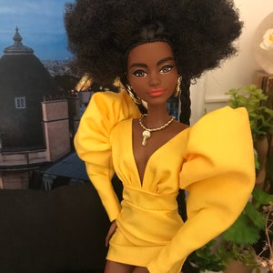 May include: A Black doll with a large afro hairstyle wearing a yellow dress with puffed sleeves and a plunging neckline. The doll is wearing gold earrings and a gold necklace with a pendant.